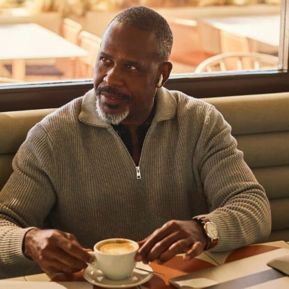 A man with a grey beard sits at a table holding a white cup of coffee.