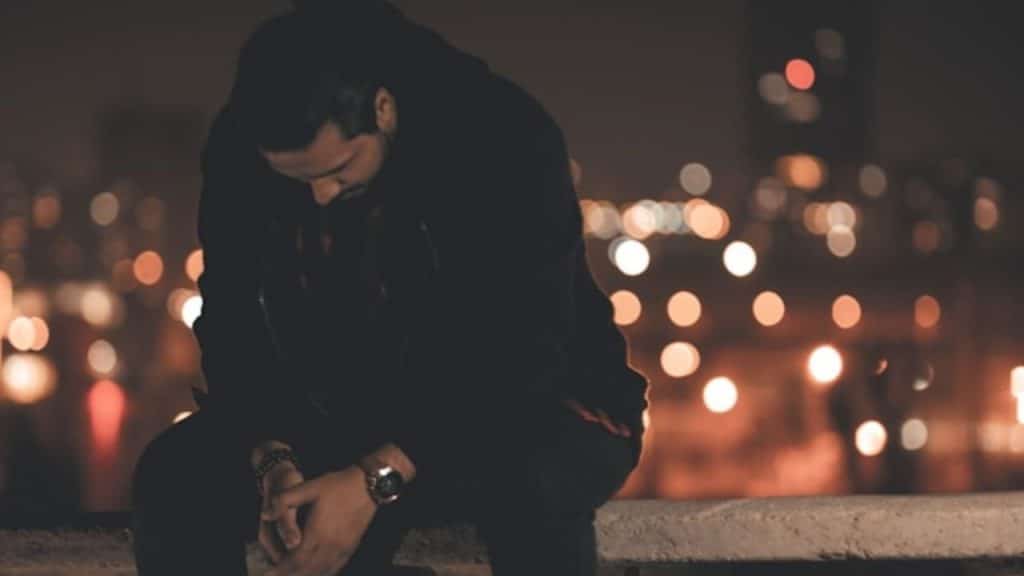 A man in dark clothing sits looking down with a blurred city lights background.