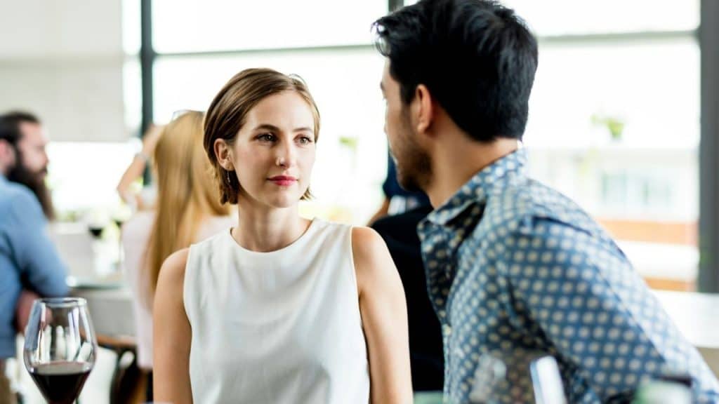 A woman in a white sleeveless top looks toward a man in a patterned shirt.