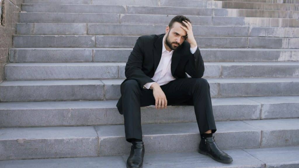 A man in a black suit sits on concrete stairs with his hand on his head.