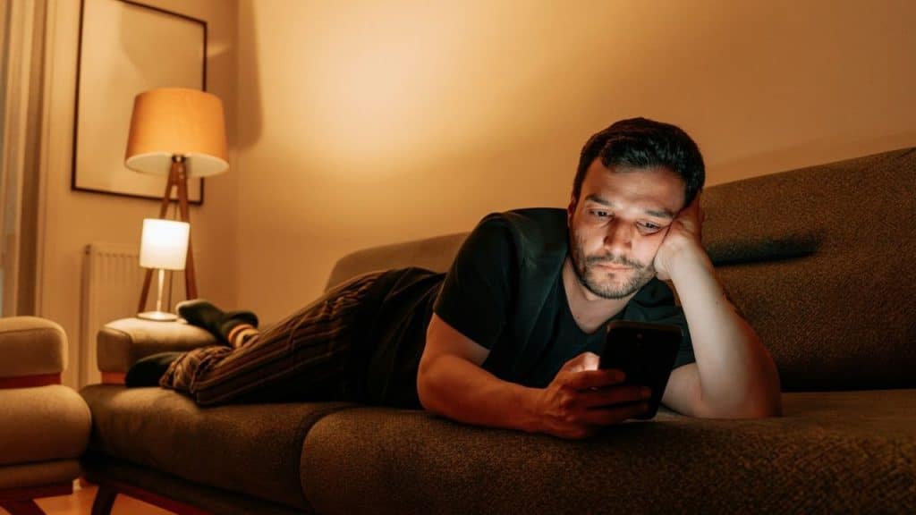 A man lies on a grey sofa in a dimly lit room looking at a smartphone.