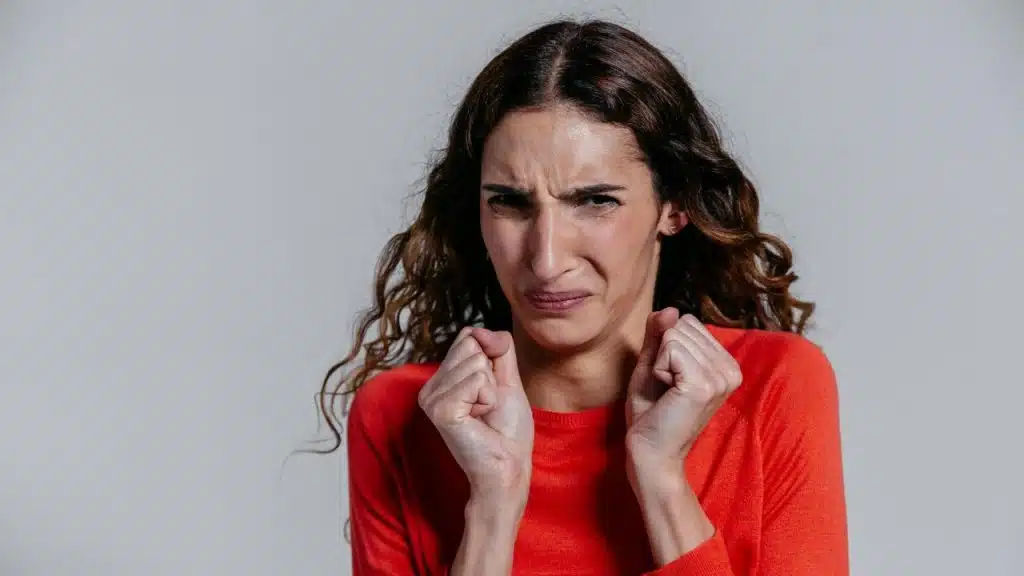 A woman with long wavy hair wears a red sweater and makes a disgusted facial expression.
