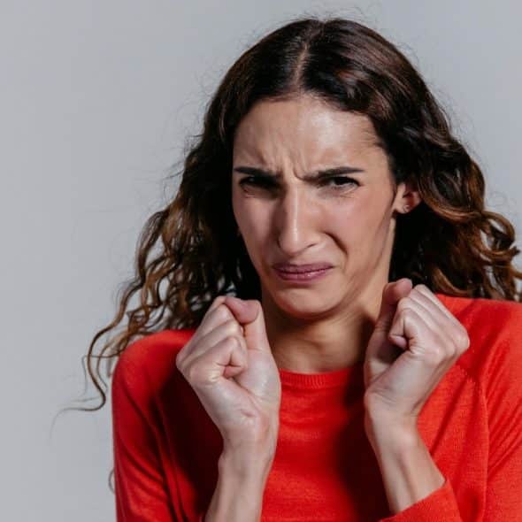 A woman with long wavy hair wears a red sweater and makes a disgusted facial expression.