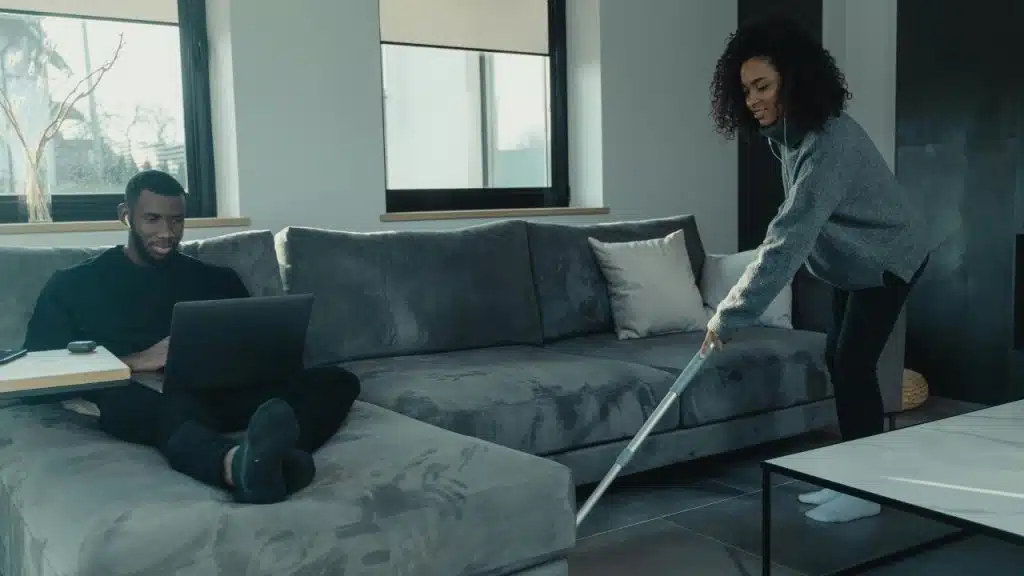 A man uses a laptop on a couch while a woman vacuums the floor.