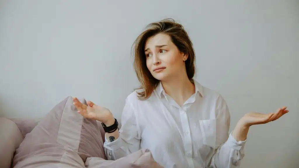 A woman in a white shirt shrugs her shoulders with her palms facing upward.