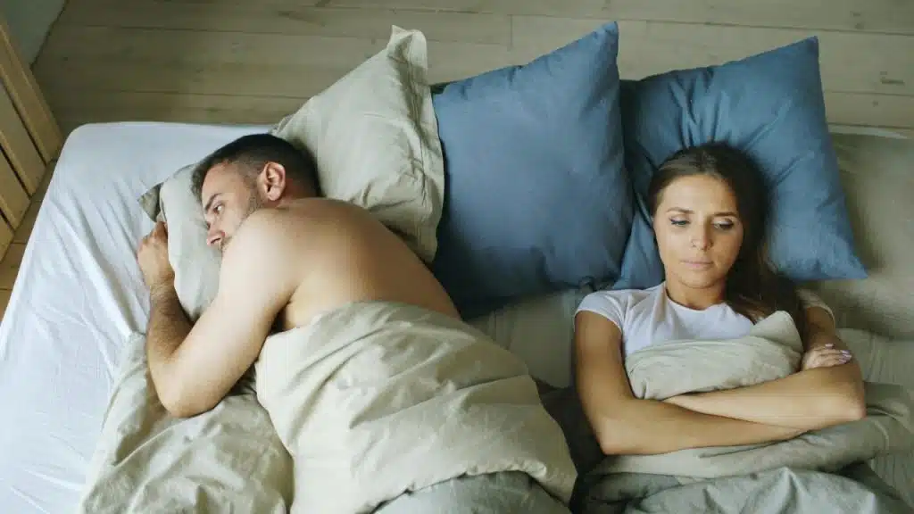 A man and woman lie in bed facing away from each other with neutral expressions.