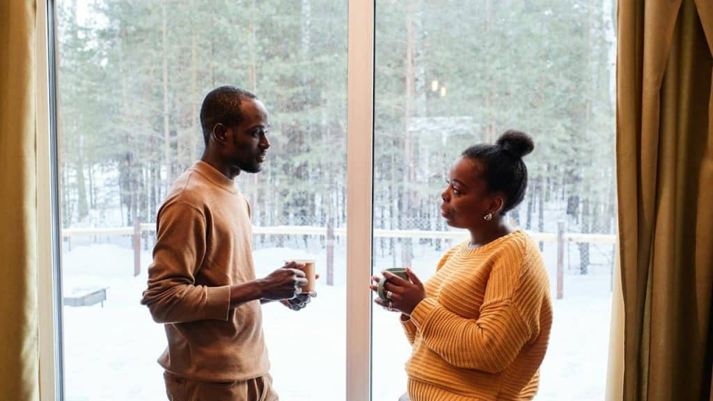 A couple having coffee and talking while standing besides a window. 