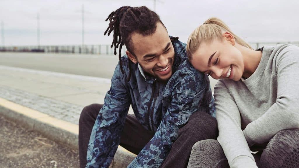 A laughing young couple sitting on a road together, taking a break from a run on an overcast day.