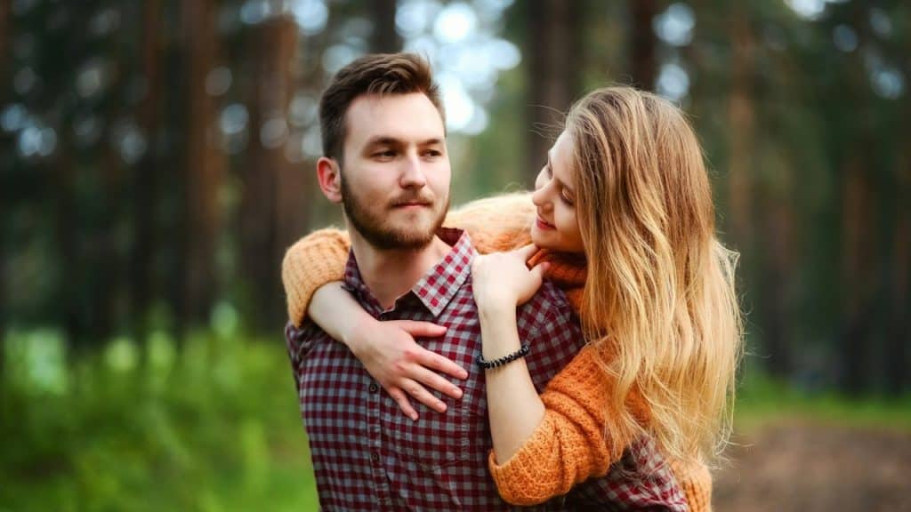 A bearded man carrying a smiling woman on his back in a forest.
