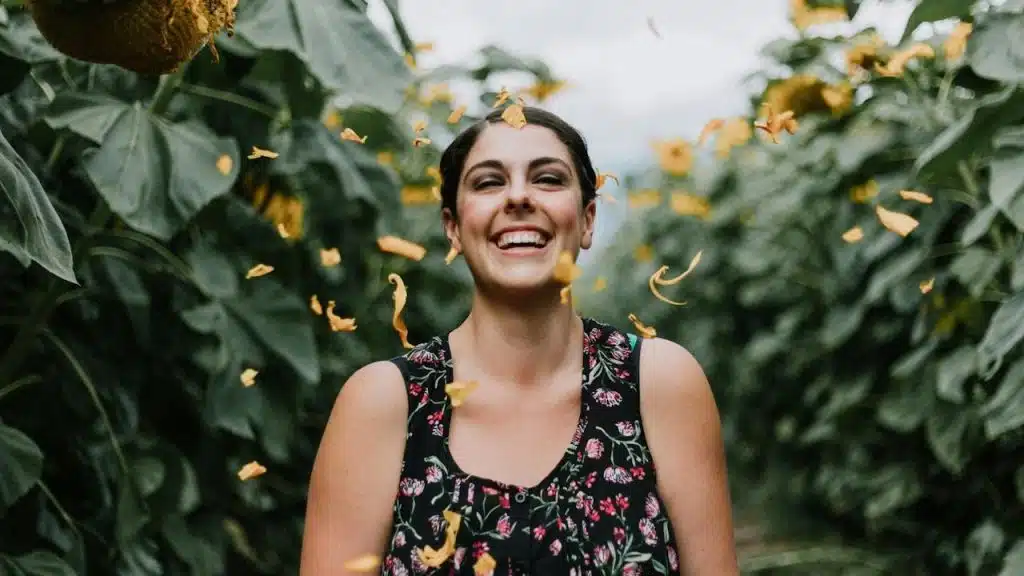 A woman in a floral top standing in an open area with big green leafy trees.