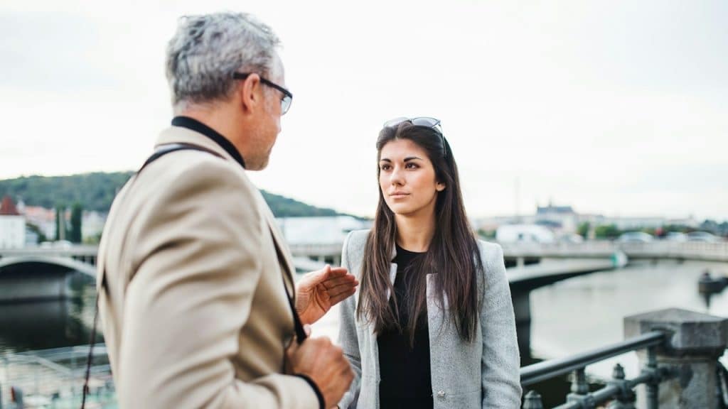 A mature man and a young woman are walking by a river in the city of Prague talking.
