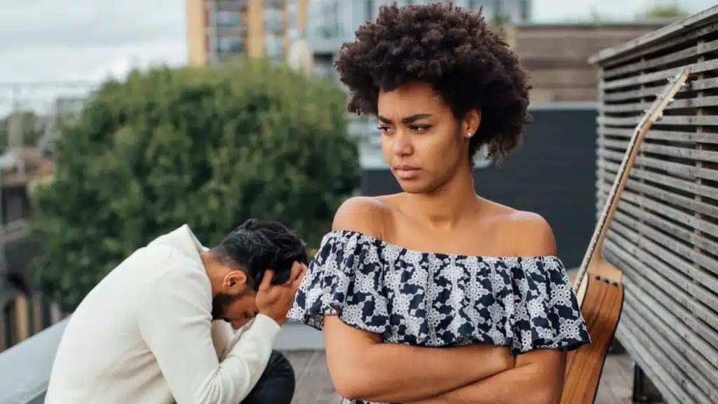 Black woman with arms crossed, kneeling and looking upset on a rooftop picnic while a man holds his head in the background