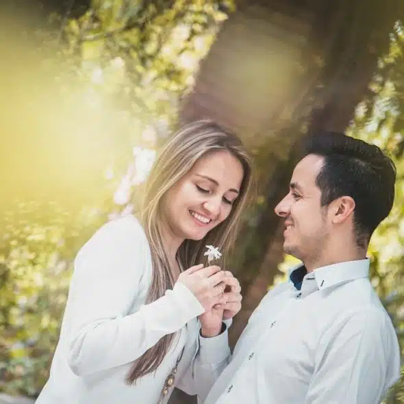 A smiling couple standing in the woods as the man hands a flower to the woman.