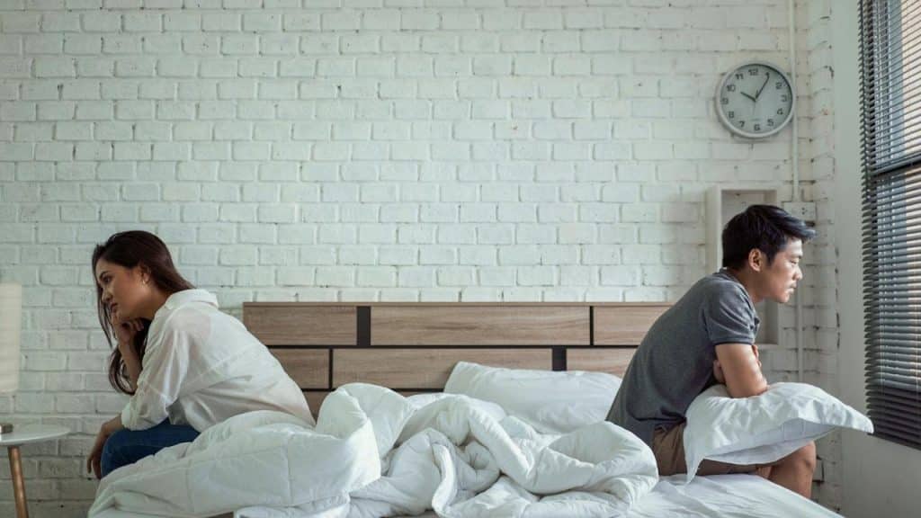 A couple sitting on separate ends of a bed facing away from each other.