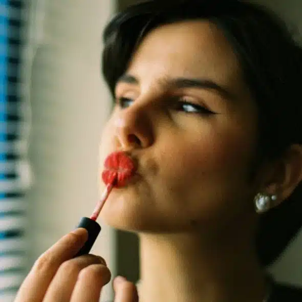 A person with short dark hair applies red liquid lipstick while looking toward a window.