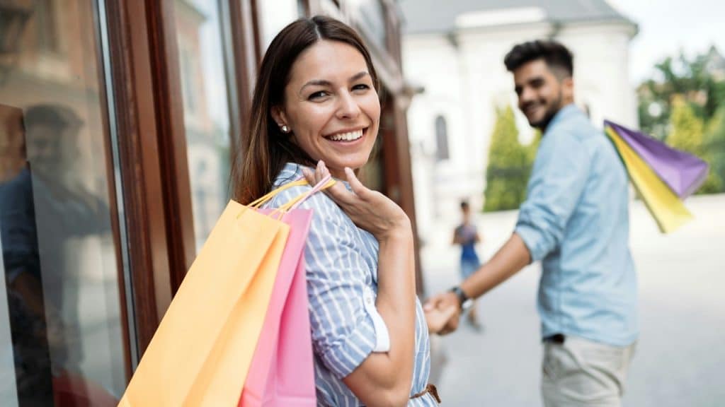 A smiling woman holds several colorful shopping bags while a man follows closely behind her.