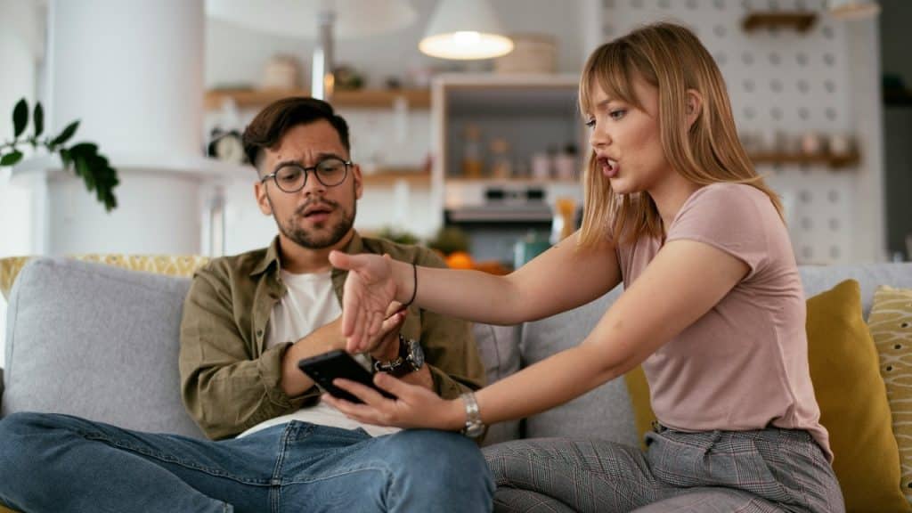 A woman gestures toward a smartphone held by a man while sitting on a couch.