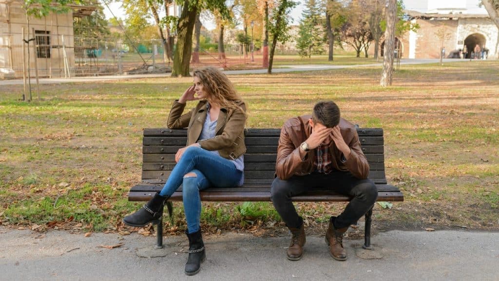 Two people sit apart on a park bench, looking in opposite directions with distressed expressions.