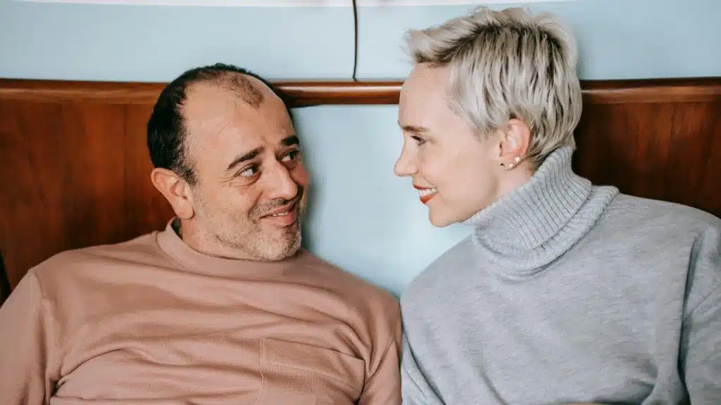 A man and a woman with short blonde hair look at each other and smile indoors.