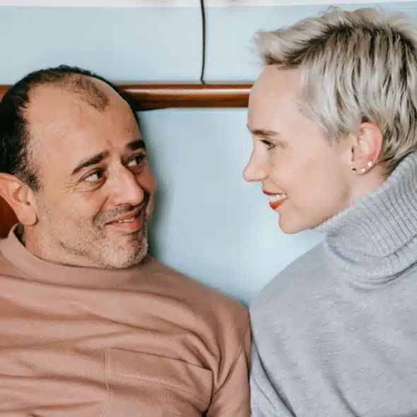 A man and a woman with short blonde hair look at each other and smile indoors.