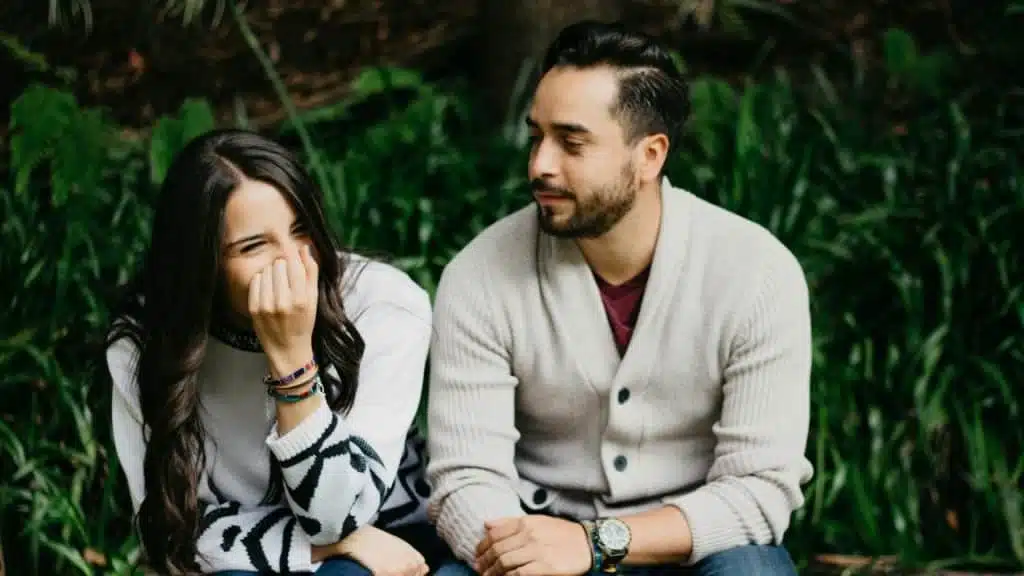 A woman laughs with her hand over her mouth while a man sits beside her outdoors.