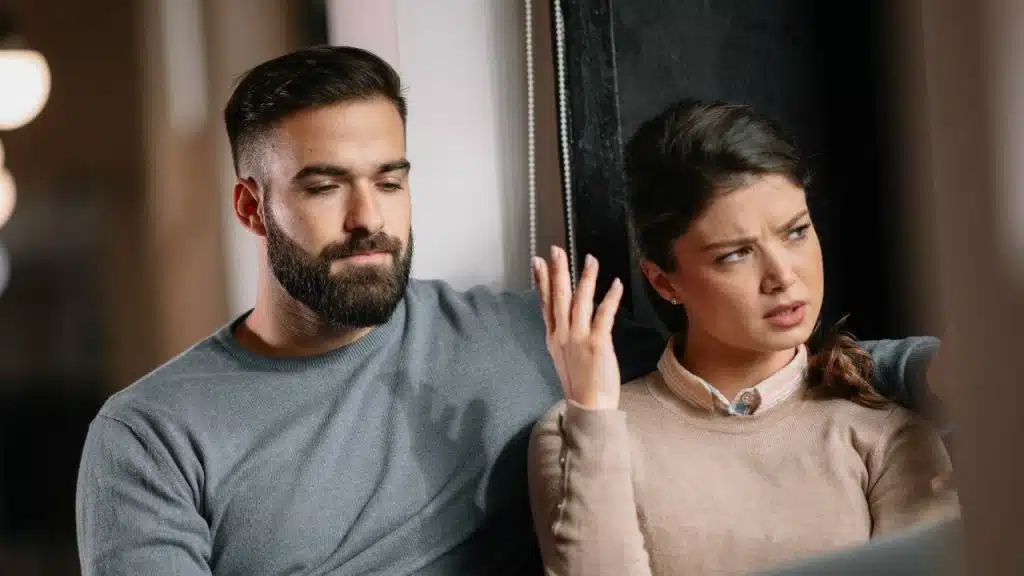 A bearded man looks down while a woman beside him looks away with an annoyed expression.