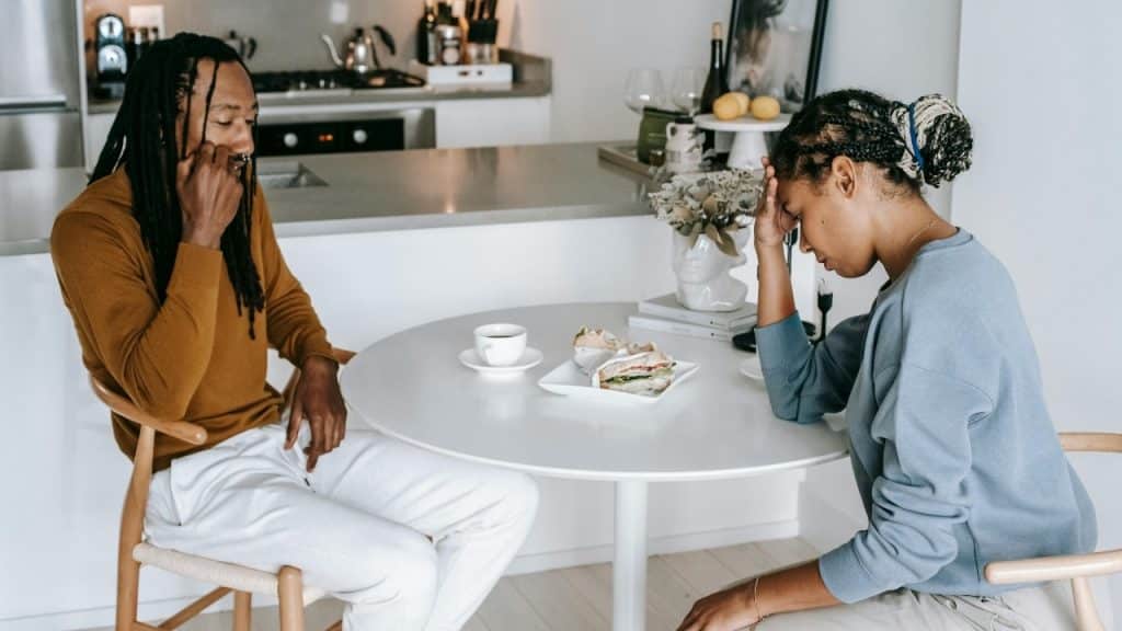 A man and woman sit at a table looking down with their hands on their heads.