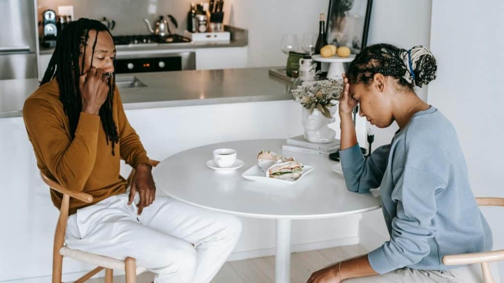 A man and woman sit at a white table with food, looking down with distressed expressions.