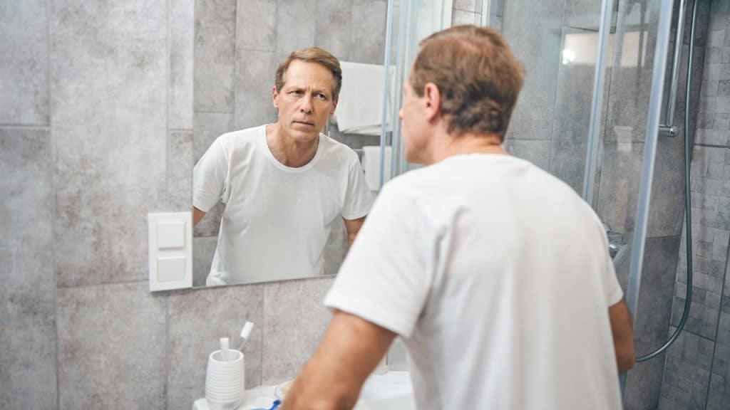 A man in a white t-shirt looks at his own reflection in a bathroom mirror.
