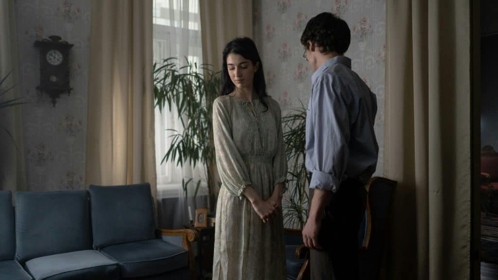 A woman in a long dress and a man stand in a dimly lit room with floral wallpaper.