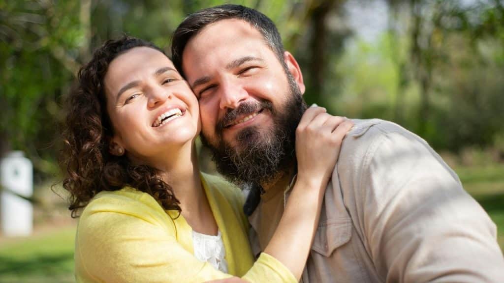 A smiling woman with curly hair and a bearded man pose closely together outdoors in sunlight.