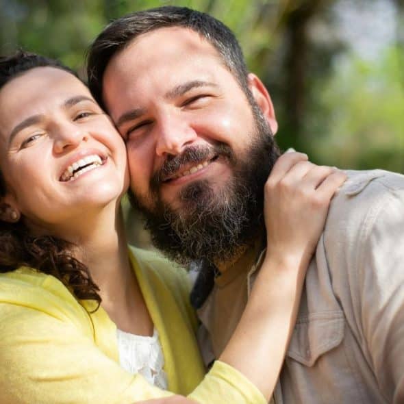 A smiling woman with curly hair and a bearded man pose closely together outdoors in sunlight.