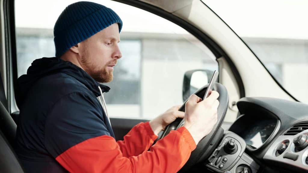 A man in a car looking at a smartphone while holding the steering wheel.