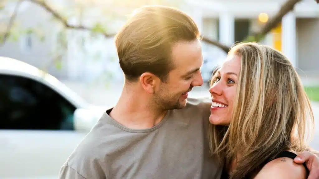 A man in a gray crew neck shirt kissing a woman in a black tank top.
