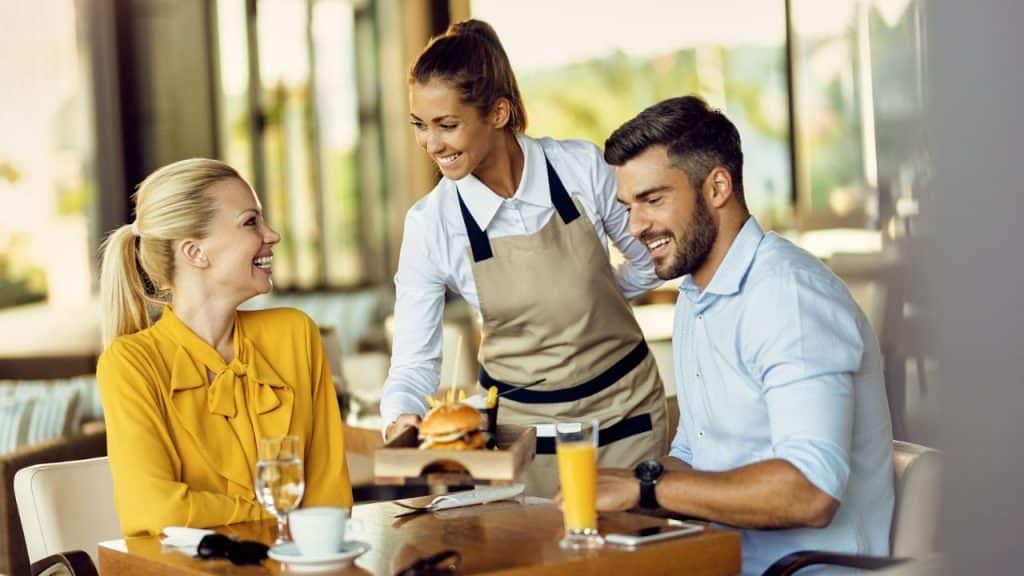 A server in an apron brings food to a smiling man and woman at a table.