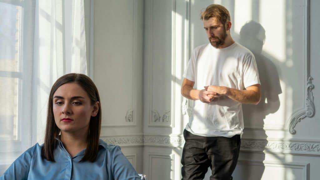 A woman looks down in the foreground while a man stands against a white wall.