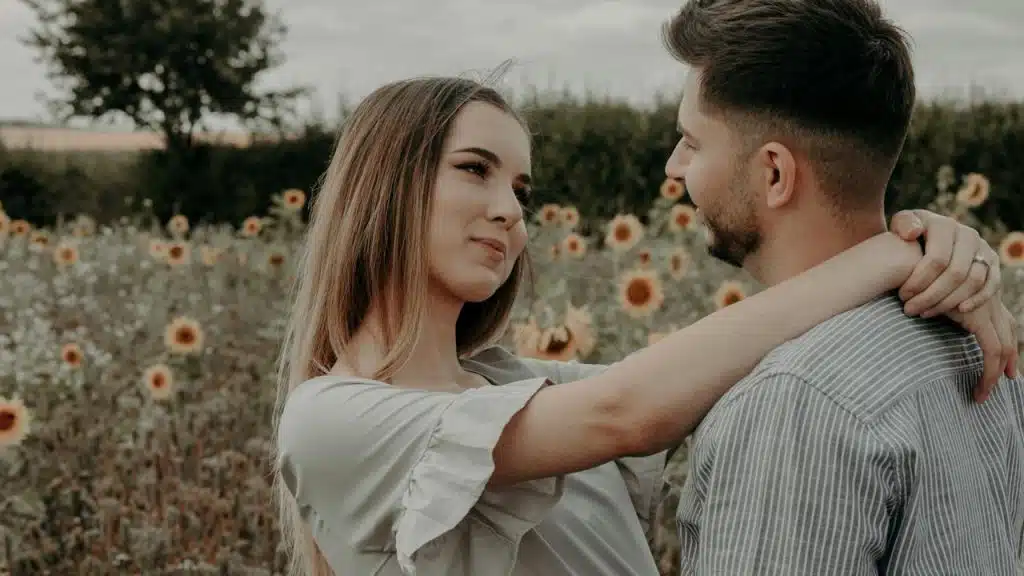 A woman staring in a man’s eyes while resting her arms on his arms as they stand in a sunflower field.