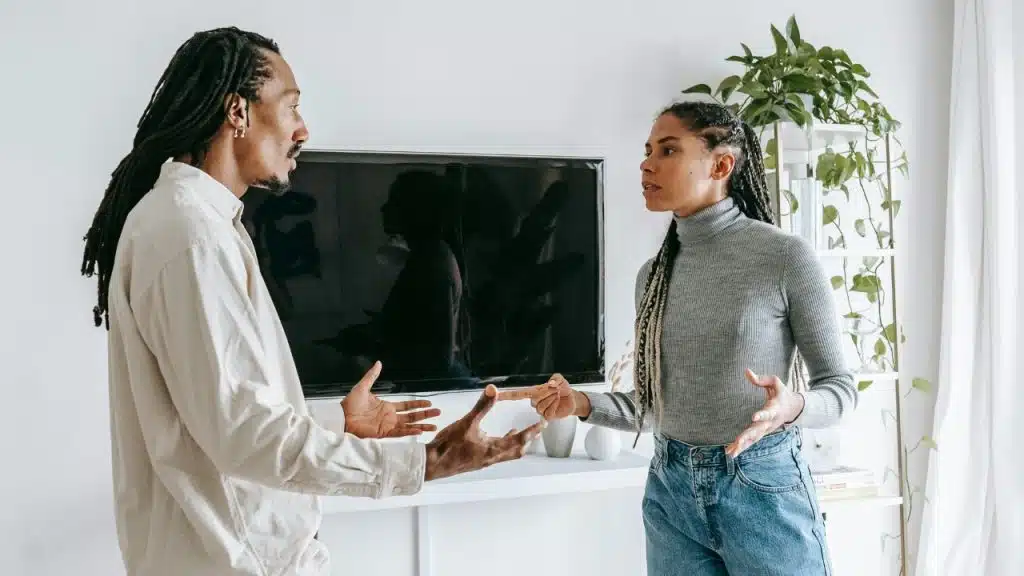 A man and woman with braids stand indoors, gesturing with their hands during a conversation.