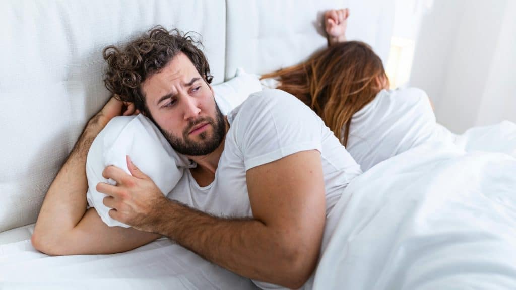 A man with curly hair lies in bed looking contemplative while a woman turns away.