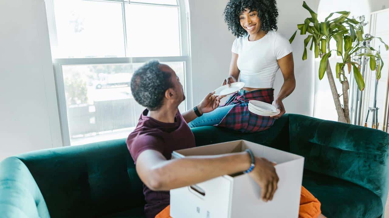 A couple unpacking dishes from a box in a bright living room.