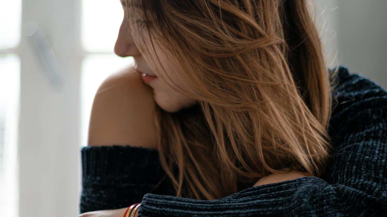 A woman with long hair looking down thoughtfully by a window.