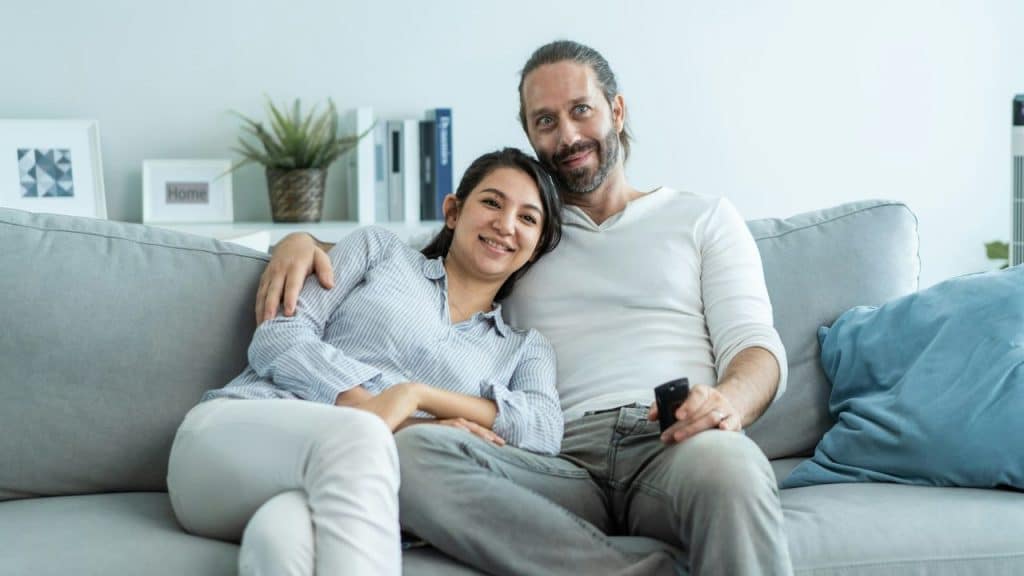 A man and woman sit together on a grey sofa, smiling while looking forward.