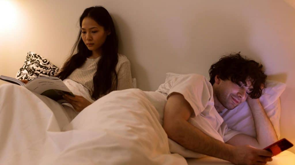 A woman reads a book in bed while a man looks at a glowing smartphone.