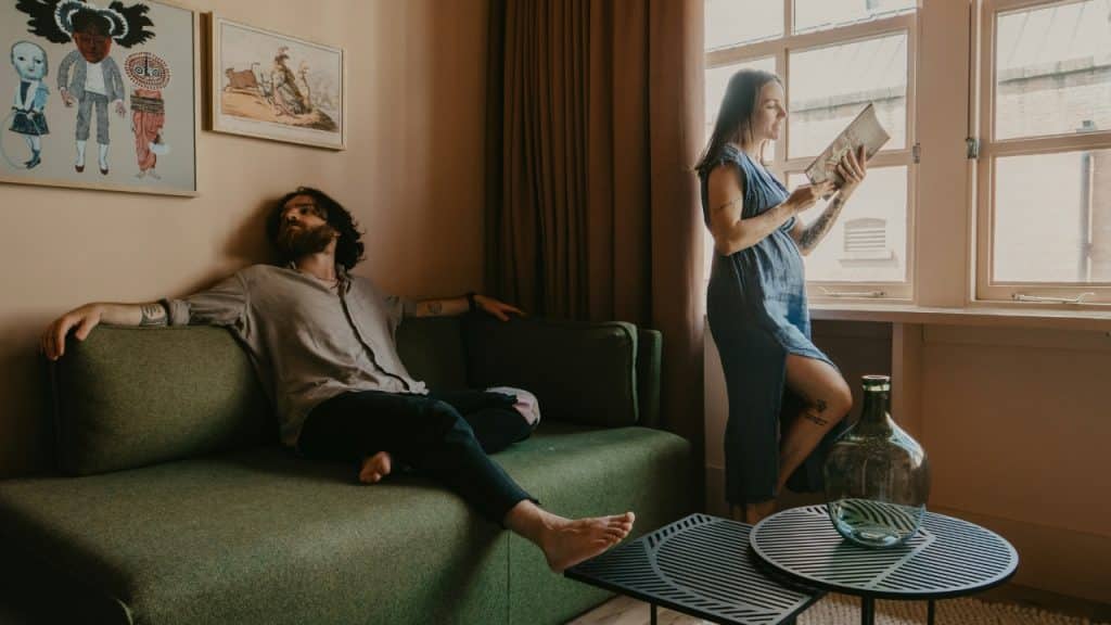 A man sits on a green sofa while a woman stands by a window reading.