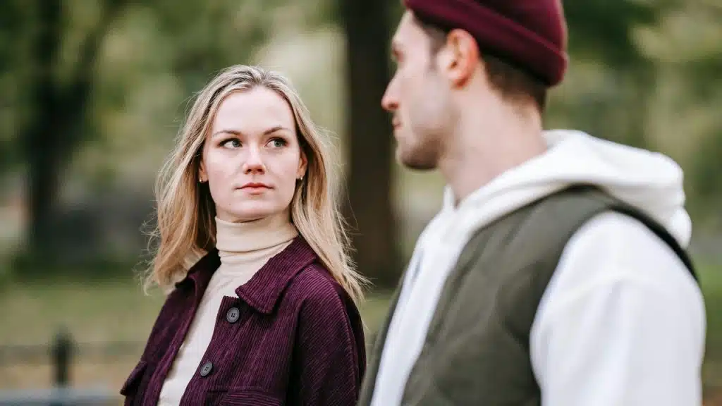 A blonde woman looks over her shoulder at a man wearing a maroon beanie outdoors.