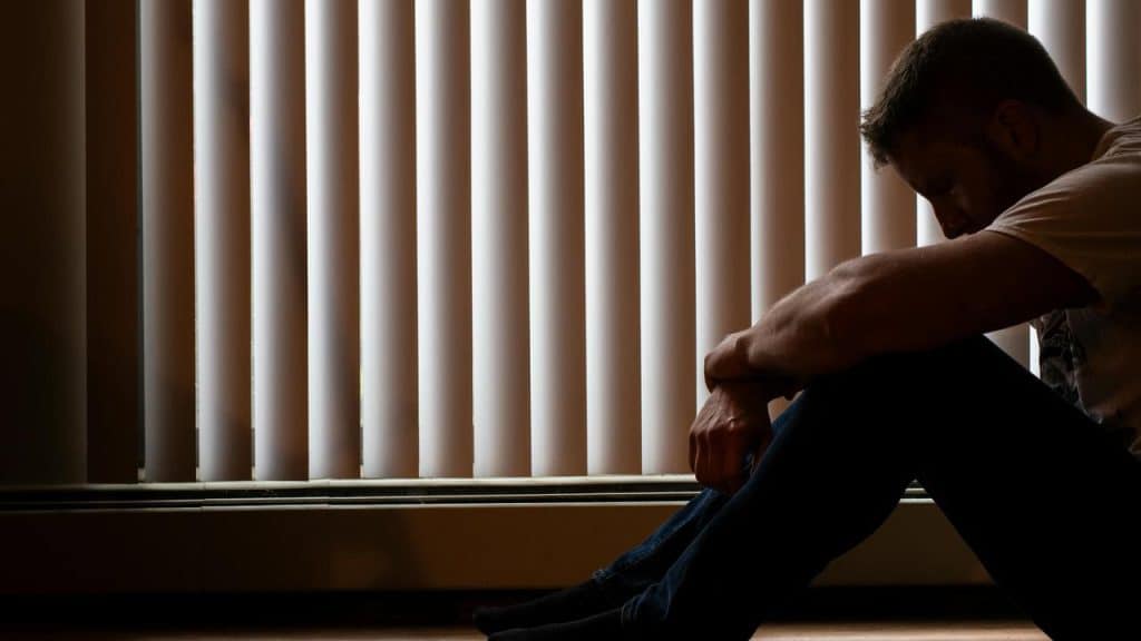 A man sitting on the floor by blinds with his head down.