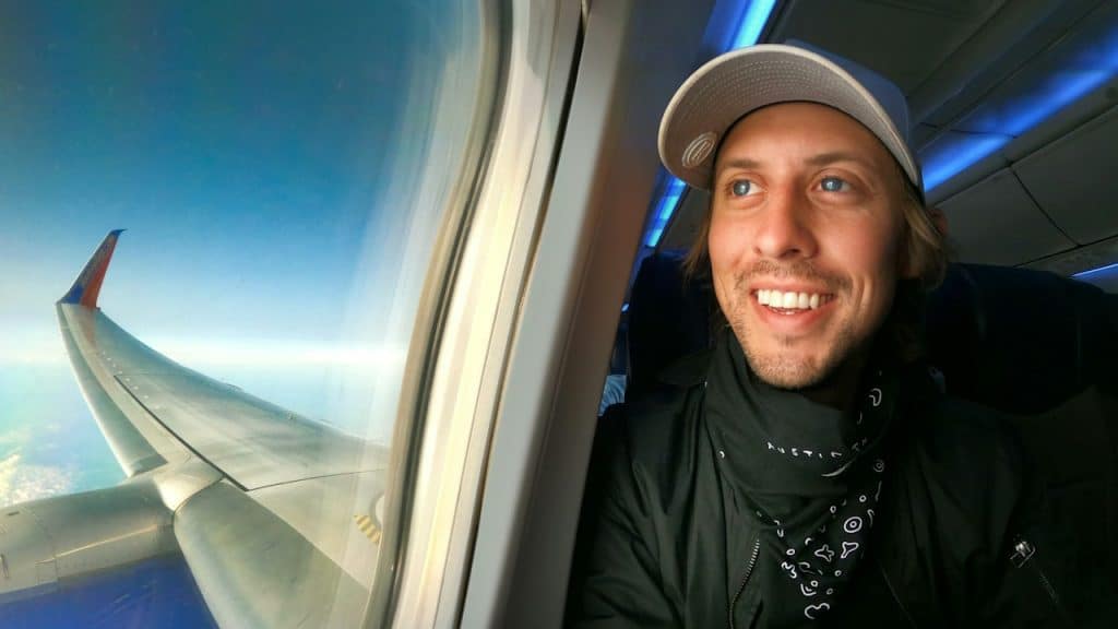 The smiling man in the black jacket and blue cap is looking out of the window of the plane.