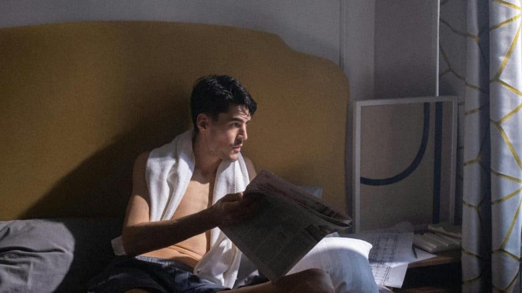 A man sitting on a bed reading a newspaper with sunlight streaming through a window.