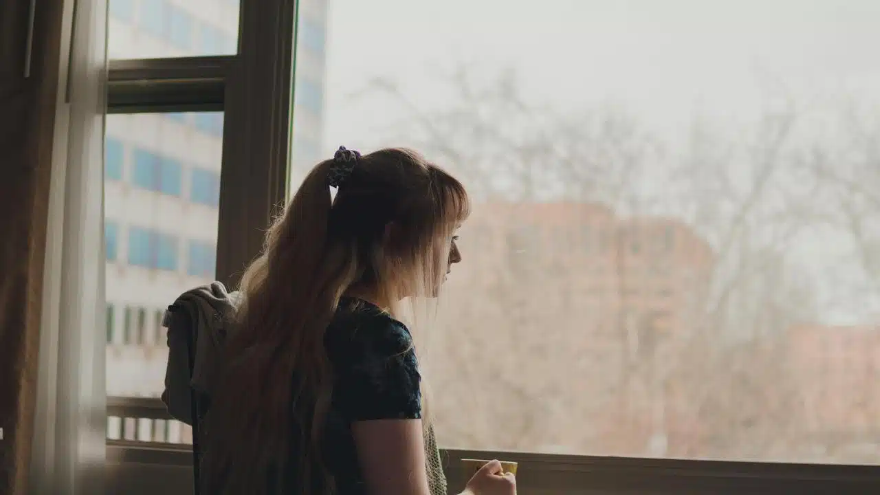 A woman standing by a window looking outside while holding a cup.
