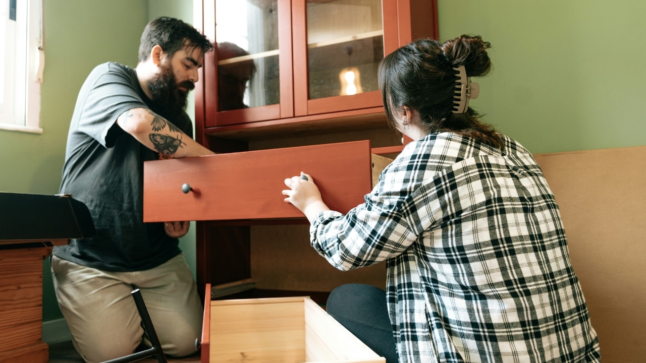 A bearded man and a woman in a plaid shirt assemble a red wooden cabinet together.